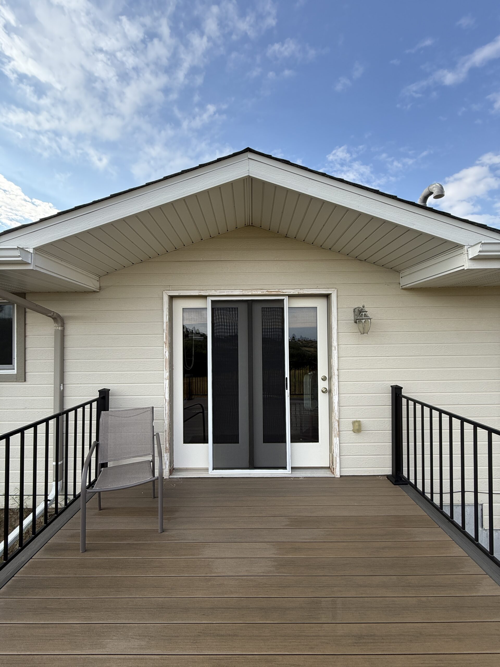 Residential Exterior Painting and Deck Staining Services Rear view of a residential house with a wooden deck, black railings, and sliding glass doors.